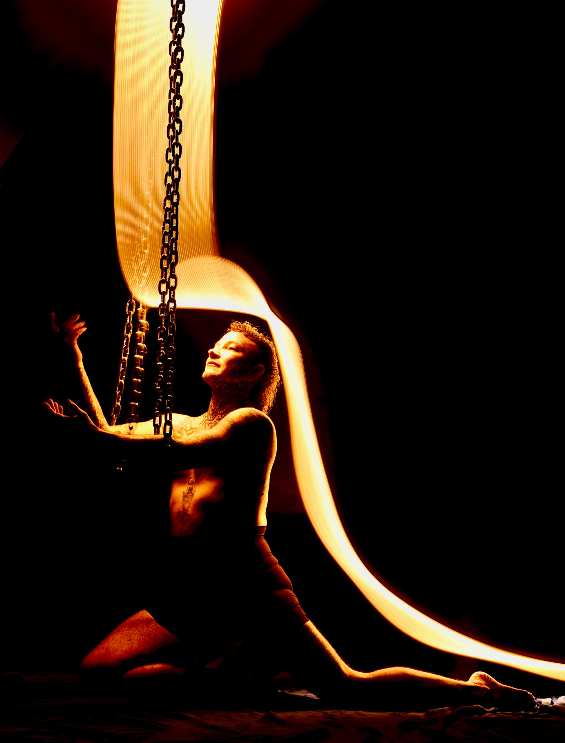 An amber-toned light-painting image where Alec is kneeling with both arms draped through the chain loops and gesturing upward.