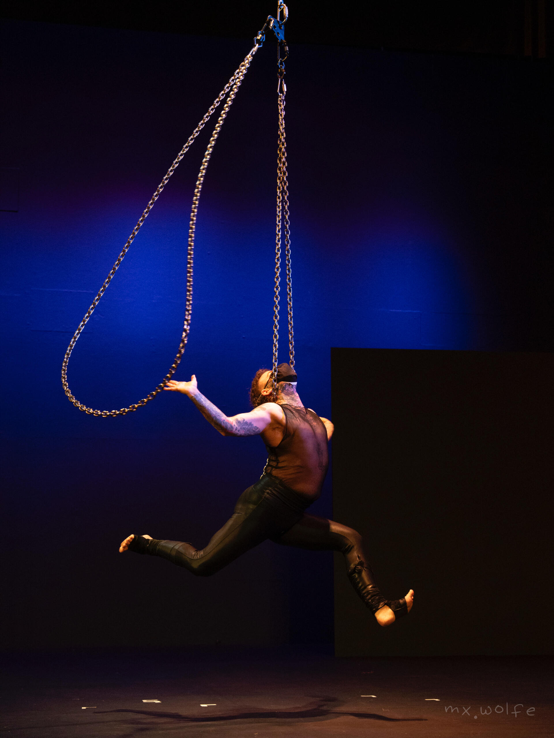 Alec suspended upright by one hip on one chain, his neck locked between the two sides and arms open, the other chain circling him.